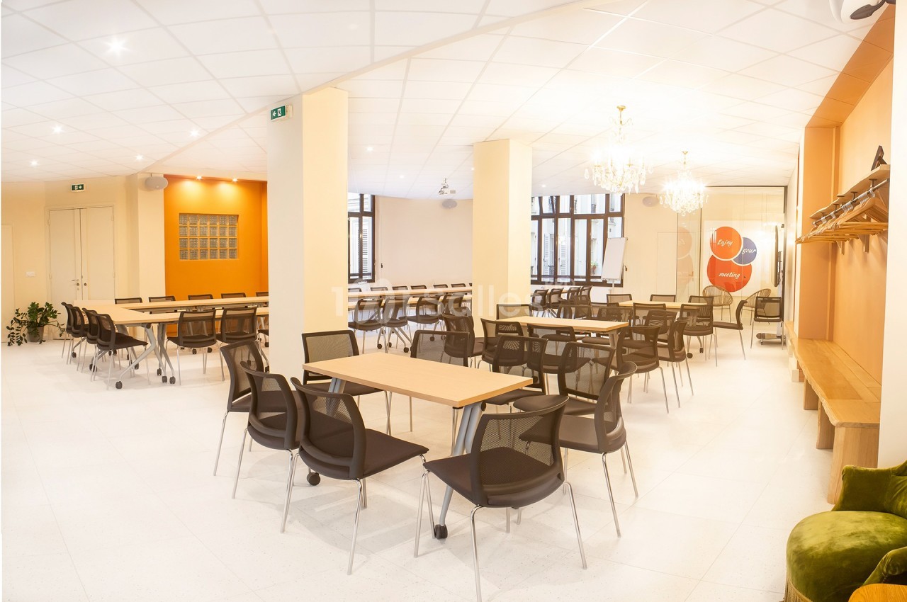 Salle lumineuse avec tables en bois, chaises noires, murs clairs et accents orange, éclairée par des lustres modernes.