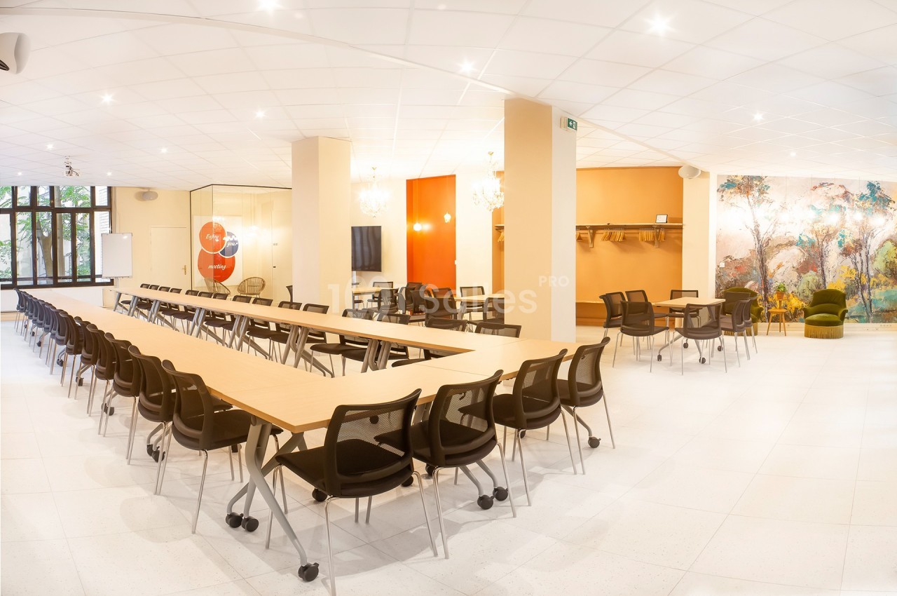 Salle lumineuse avec de longues tables en bois, des chaises noires, un mur décoré et un espace détente en arrière-plan.