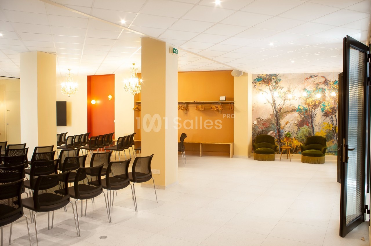 Salle lumineuse avec rangées de chaises noires, espace de vestiaire, murs colorés et fresque murale décorative.