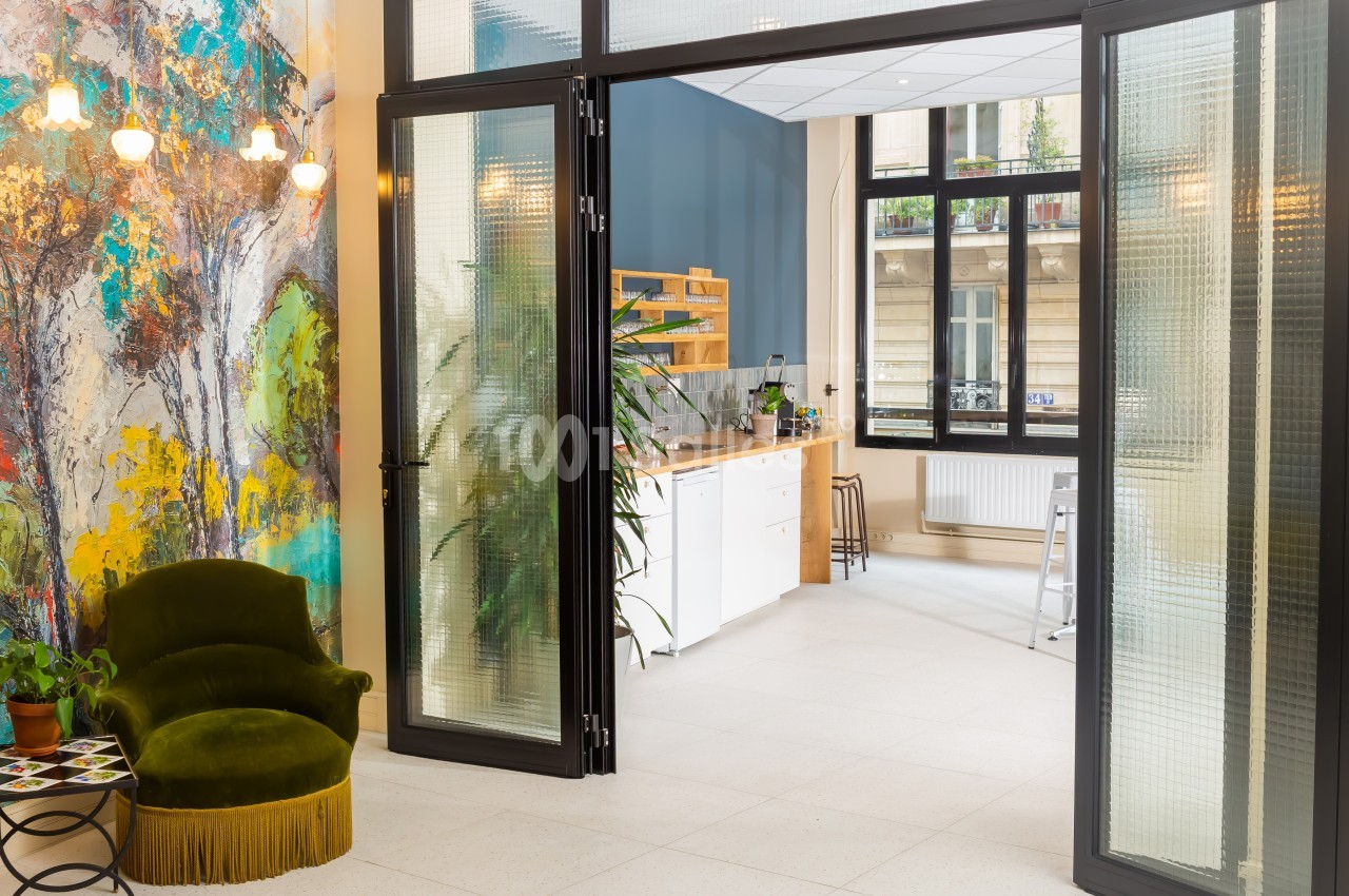 Espace lumineux avec fauteuils verts, mur coloré, cuisine ouverte et grandes fenêtres donnant sur l'extérieur.