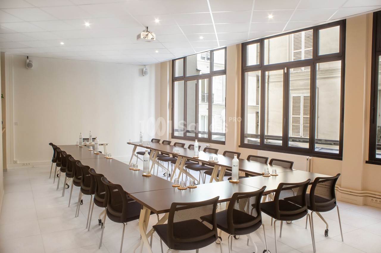 Salle de réunion lumineuse avec de grandes fenêtres, des tables disposées en U et des chaises noires.