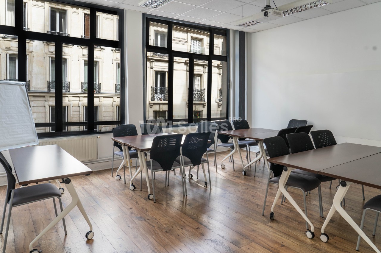Salle de réunion lumineuse avec tables en bois, chaises noires et grandes fenêtres donnant sur des immeubles.