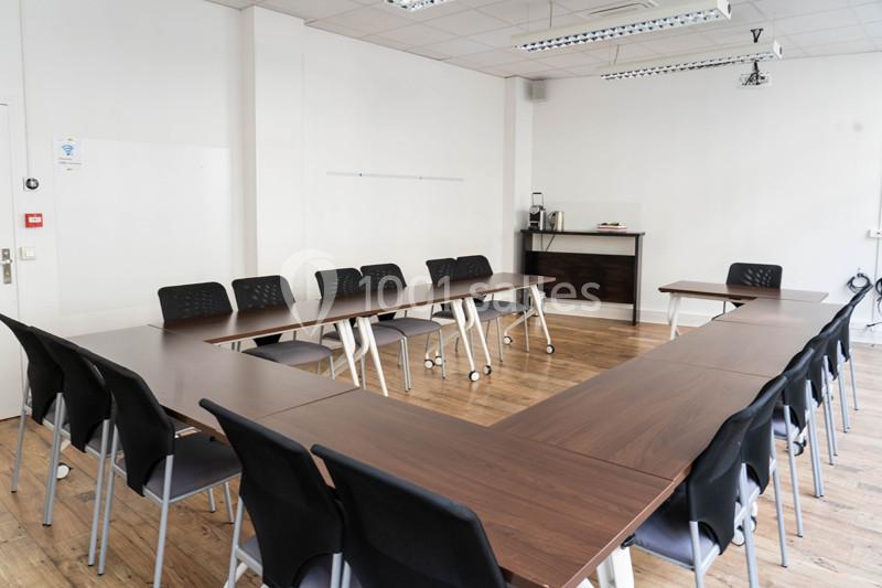 Salle de réunion lumineuse avec tables en disposition en U, chaises noires et un coin avec cafetière en arrière-plan.