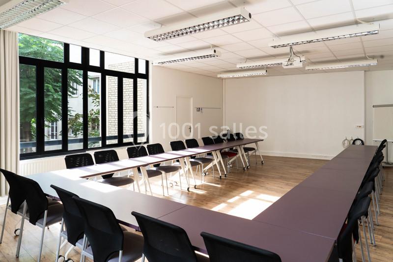 Salle de réunion lumineuse avec tables disposées en U, chaises noires et grandes fenêtres donnant sur un espace arboré.
