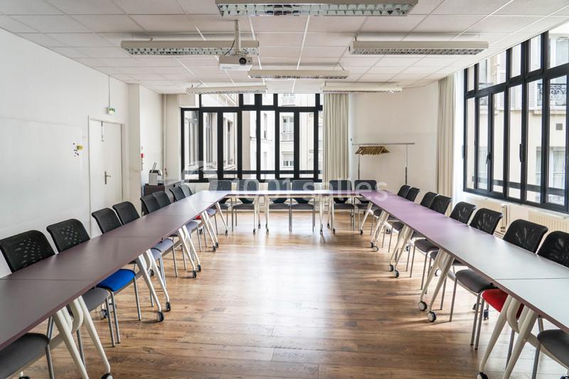 Salle de réunion lumineuse avec tables disposées en U, chaises noires et grandes fenêtres donnant sur l'extérieur.