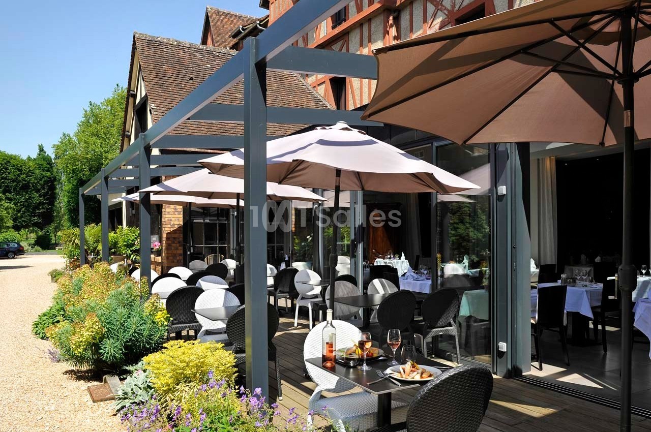 Terrasse d'un restaurant avec tables dressées, chaises en osier et parasols, entourée de végétation et d'un bâtiment en…