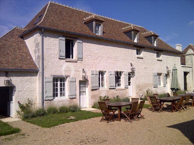 Façade d'une maison en pierre avec volets bleus, toit en tuiles et tables en bois dans une cour gravillonnée.