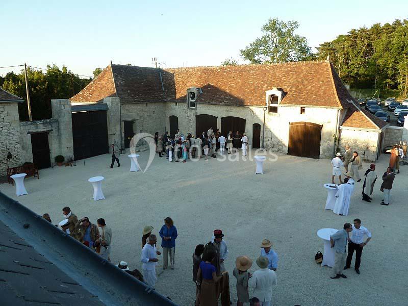 Groupe de personnes rassemblées dans une cour extérieure devant un bâtiment en pierre avec toit en tuiles.