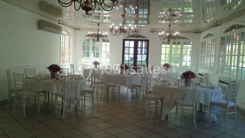 Salle lumineuse avec tables rondes dressées, chaises blanches et décorations florales, entourée de grandes fenêtres.