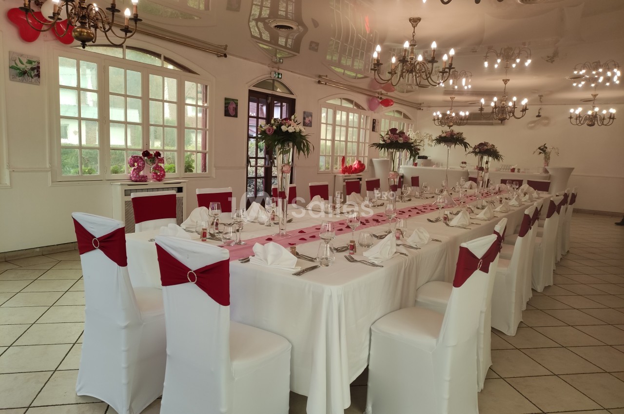 Salle de réception décorée pour un événement, avec une longue table dressée, chaises blanches et nœuds rouges.
