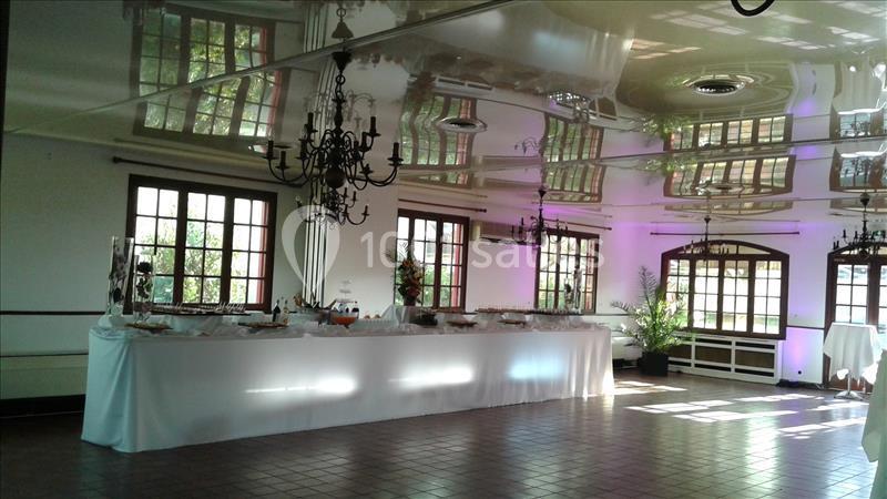 Salle lumineuse avec grandes fenêtres, plafond réfléchissant, lustres noirs et table de buffet dressée.