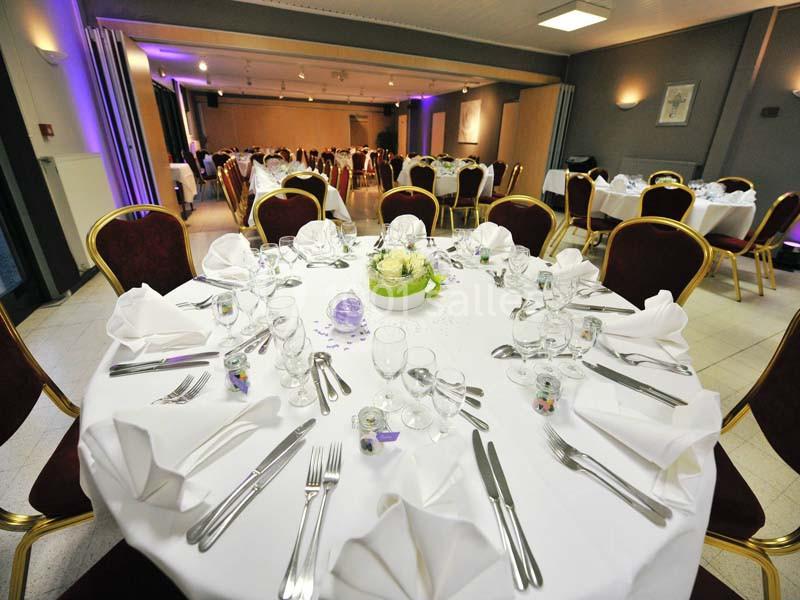 Salle de réception avec tables rondes dressées, nappes blanches, serviettes pliées et décoration centrale florale.