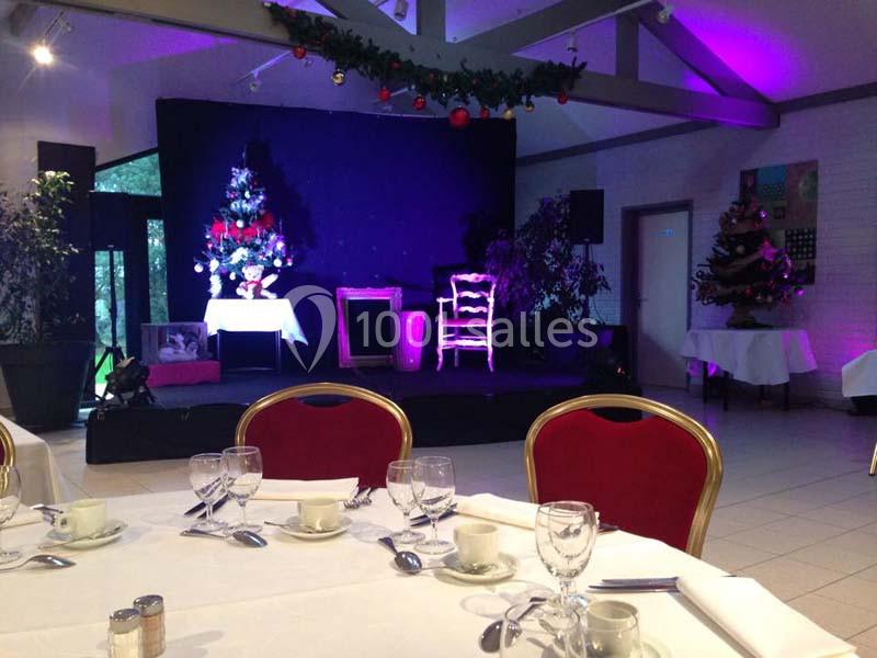 Scène décorée avec un sapin de Noël illuminé, entourée de chaises rouges et de tables dressées dans une salle festive.