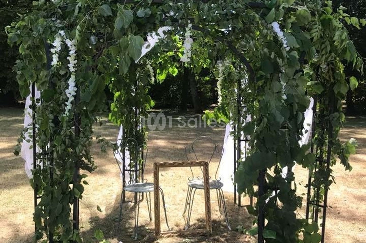 Tonnelle décorée de feuillage et de fleurs blanches, abritant deux chaises et une petite table en métal, dans un jardin.