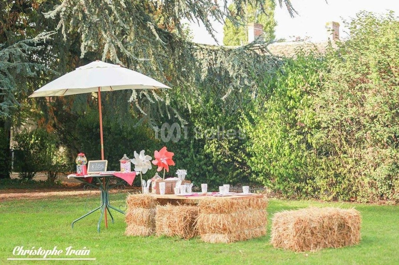 Table basse en bottes de paille avec décoration champêtre, sous un parasol blanc dans un jardin verdoyant.