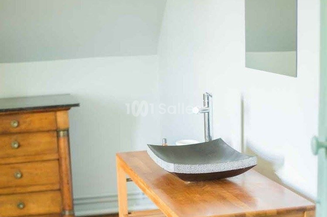 Lavabo moderne en pierre sur une table en bois, avec un miroir et une commode en arrière-plan dans une pièce lumineuse.