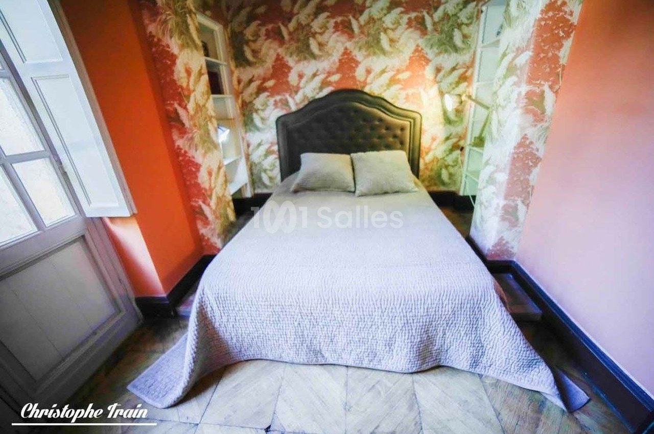 Lit double avec tête de lit capitonnée dans une chambre aux murs ornés de papier peint floral et éclairée par deux fenêtres.