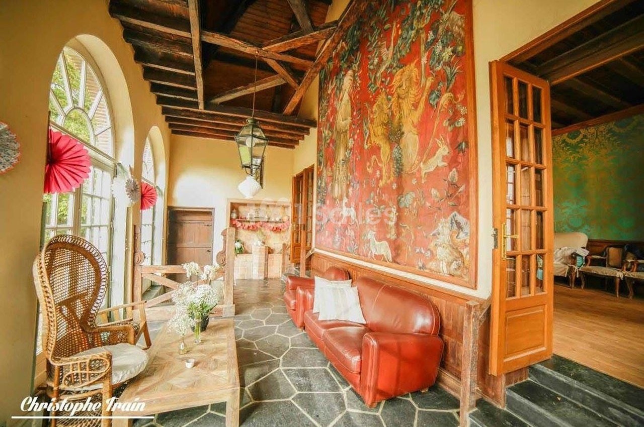 Salon lumineux avec canapé en cuir rouge, fauteuil en osier, grandes fenêtres et fresque murale colorée.