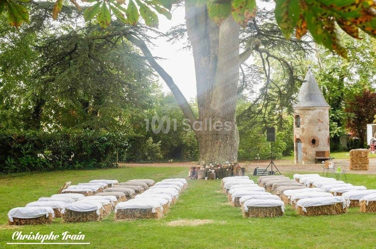 Chaises en bottes de paille recouvertes de tissu blanc disposées en extérieur sous un grand arbre, près d'une tour en pierre.
