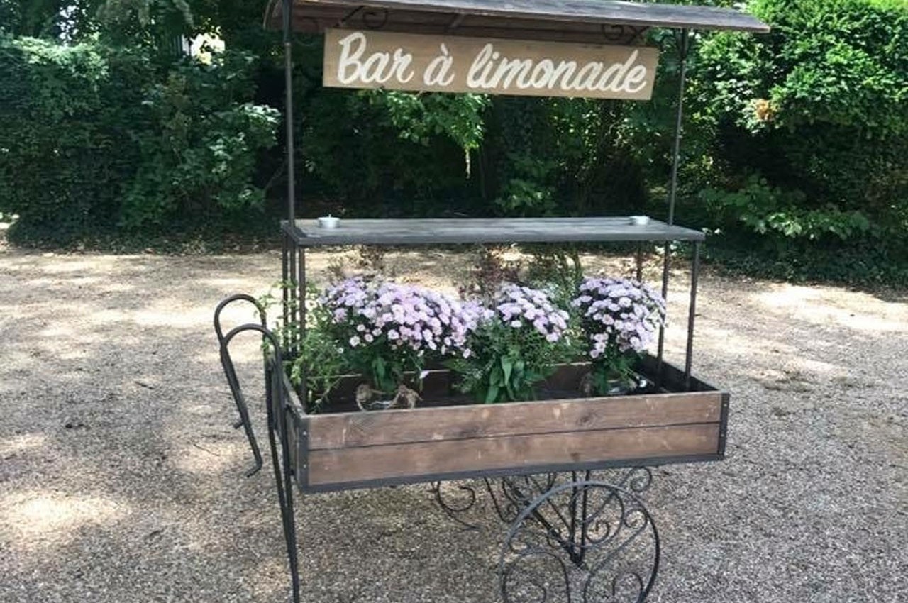 Chariot en bois et métal avec un panneau ’Bar à limonade’, décoré de fleurs violettes, placé sur un sol gravillonné.