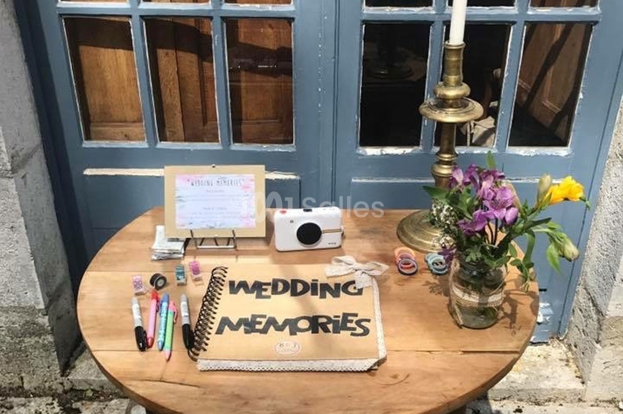 Table ronde en bois avec un album ’Wedding Memories’, des stylos, un appareil photo, des fleurs et une bougie devant une…