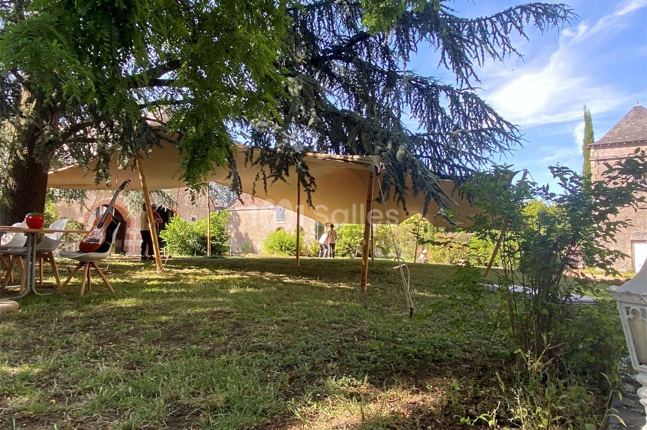 Grande tente beige installée dans un jardin ombragé, entourée de chaises et de tables sous des arbres.