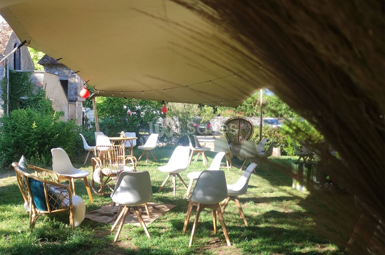 Espace extérieur aménagé avec des chaises et fauteuils sous une tente, entouré de verdure et de lumière naturelle.