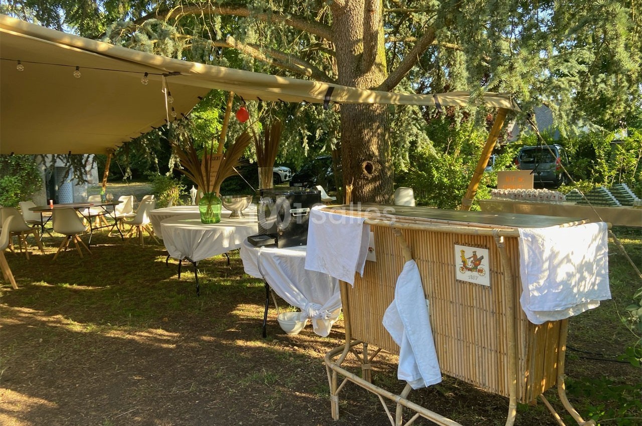 Espace extérieur ombragé avec tables, chaises et un comptoir en bambou sous un grand arbre.