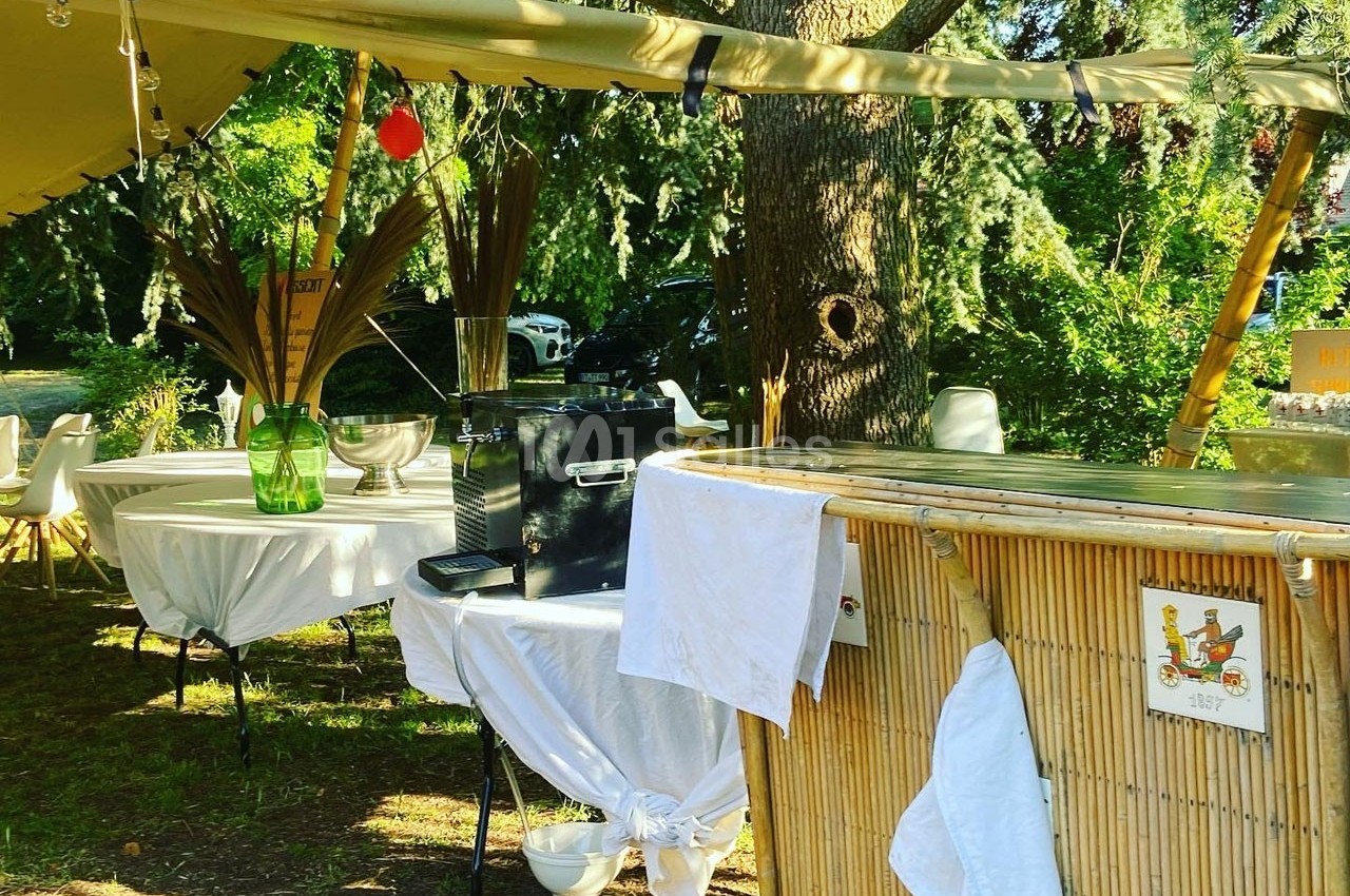 Stand extérieur avec bar en bambou, tables décorées et matériel sous une tente, entouré de végétation.