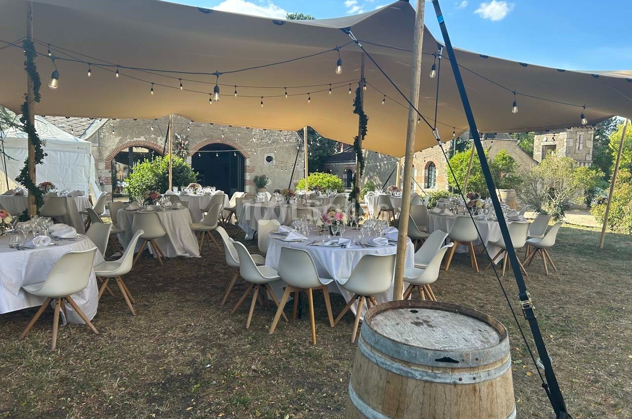 Tables dressées sous une tente beige avec guirlandes lumineuses, dans un jardin près de bâtiments en pierre.