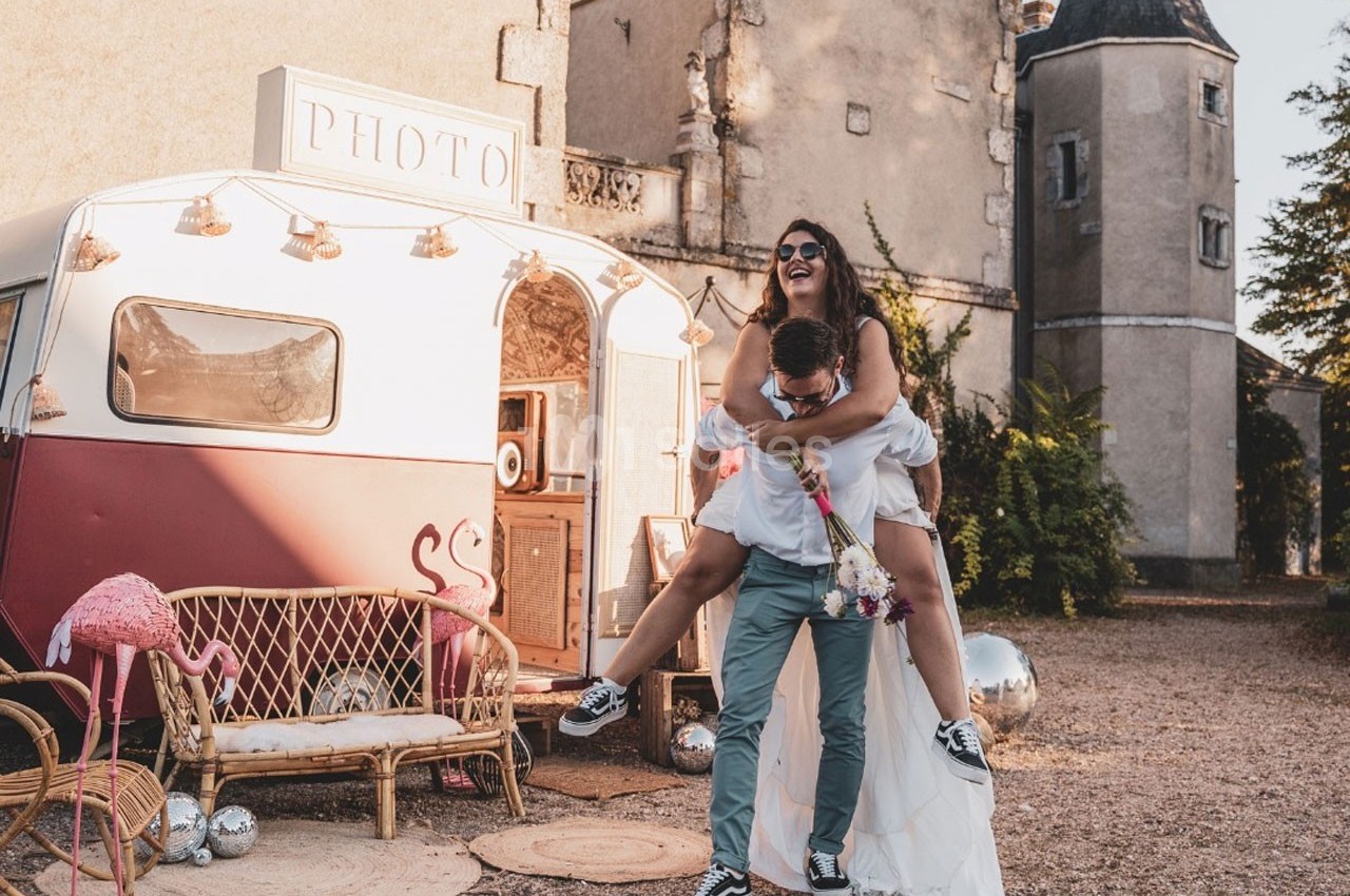 Un couple en tenue de mariage s'amuse devant une caravane rétro transformée en photobooth, près d'un bâtiment ancien.