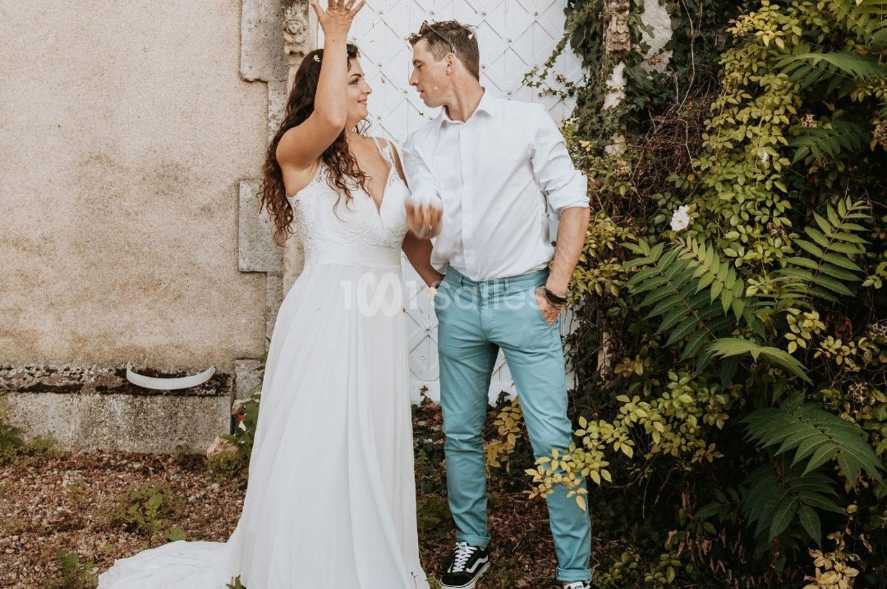 Une femme en robe blanche et un homme en chemise discutent devant un mur clair entouré de végétation.