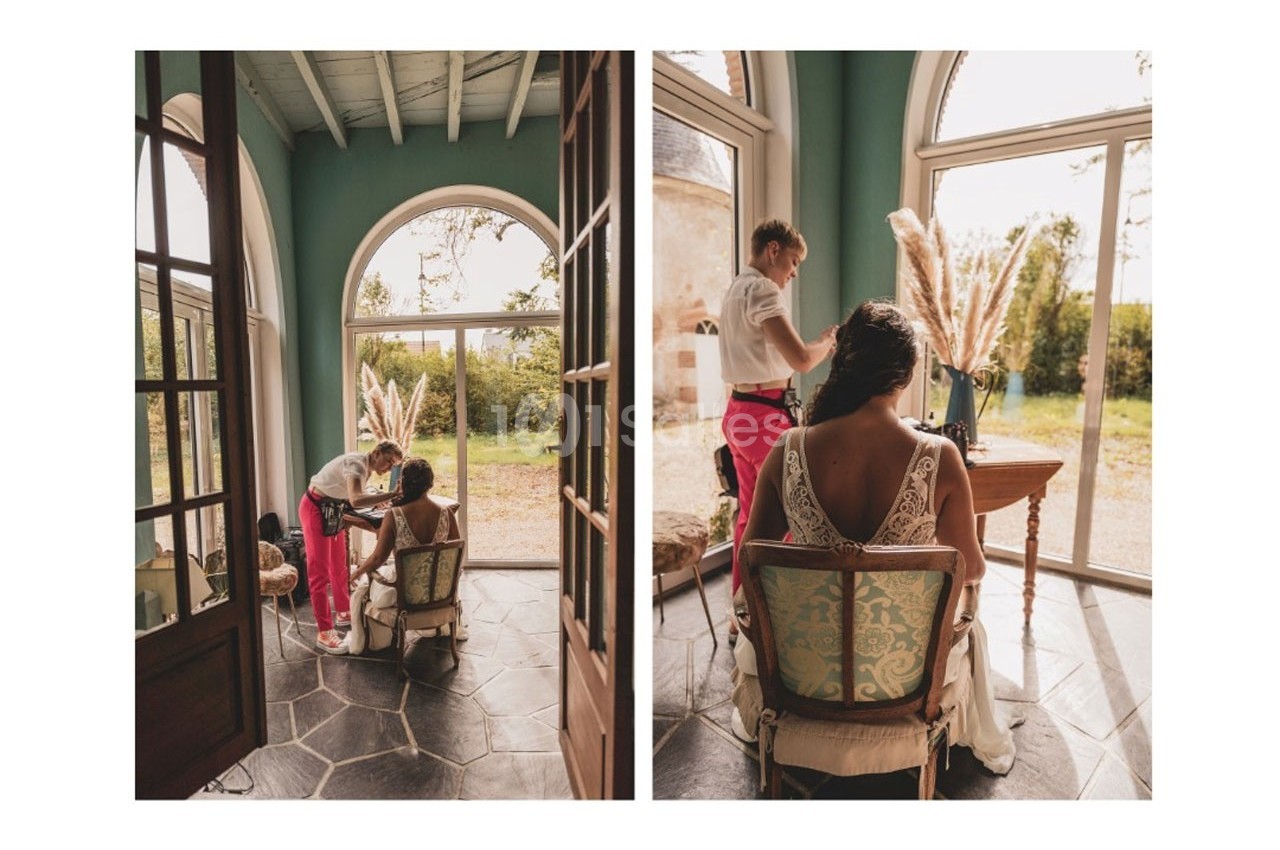 Une coiffeuse prépare les cheveux d'une femme en robe blanche, assise près d'une grande fenêtre lumineuse.