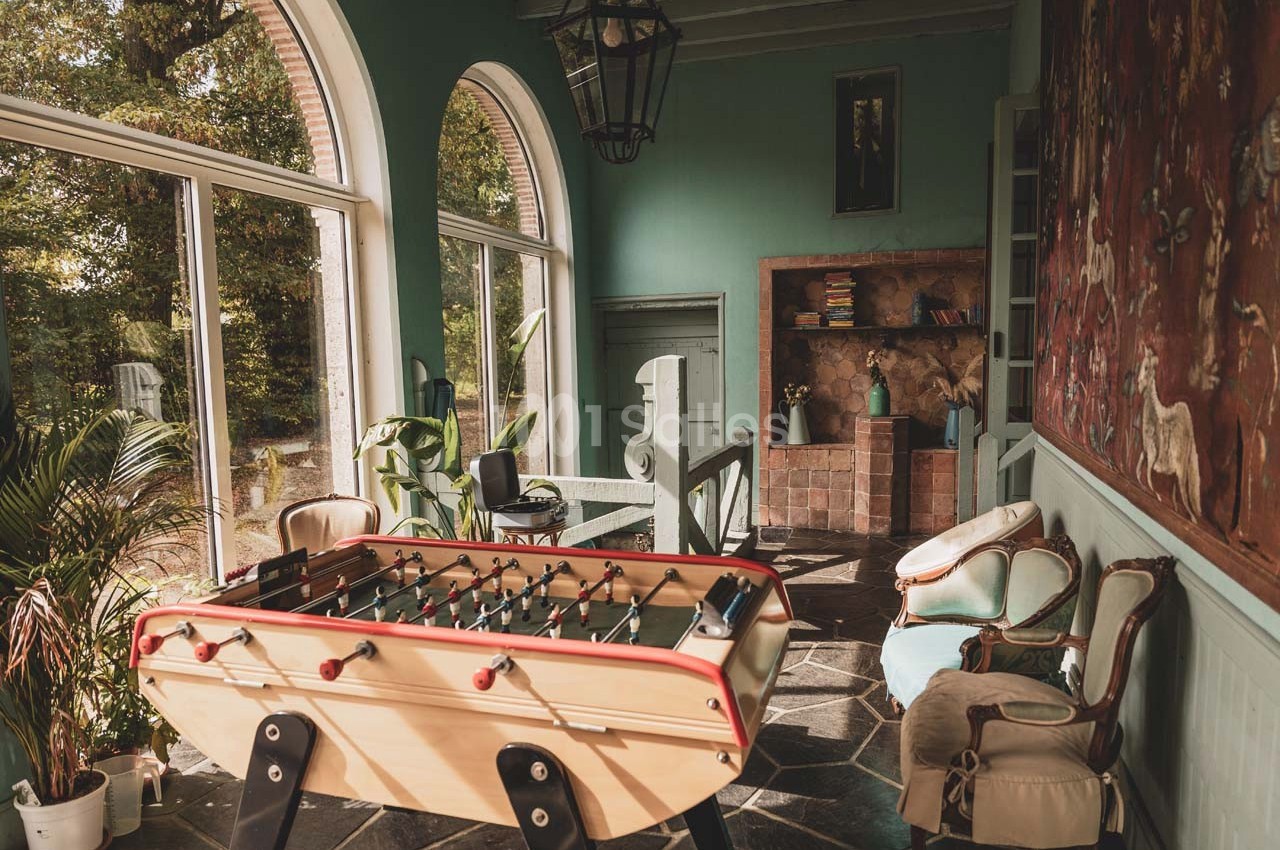 Salle lumineuse avec baby-foot, fauteuils anciens, plantes et grande baie vitrée donnant sur un jardin verdoyant.