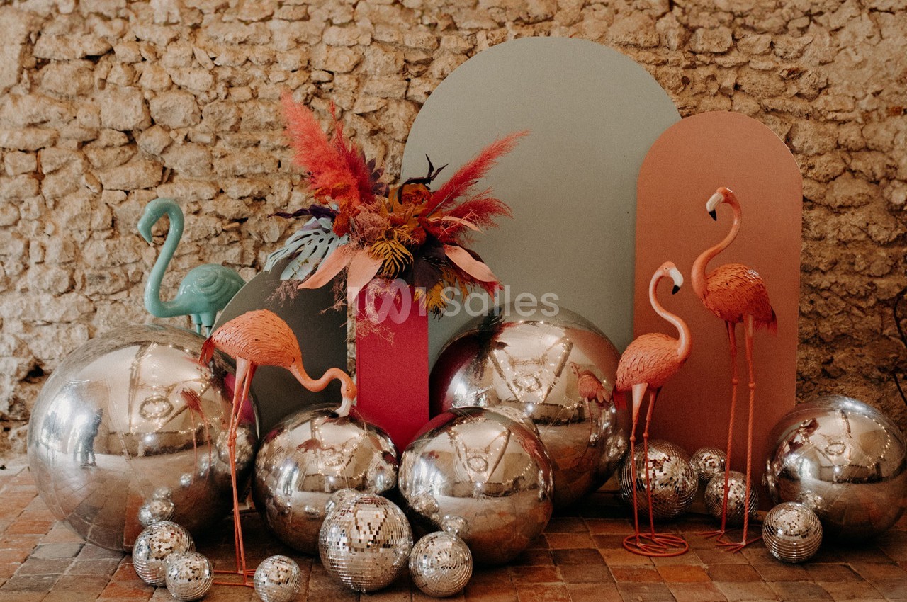 Décoration festive avec flamants roses, boules miroir et fleurs colorées devant un mur en pierre.