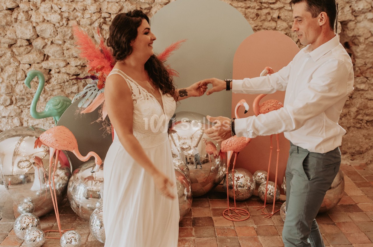 Un couple danse joyeusement dans un décor coloré avec des flamants roses, des ballons et un mur en pierre.