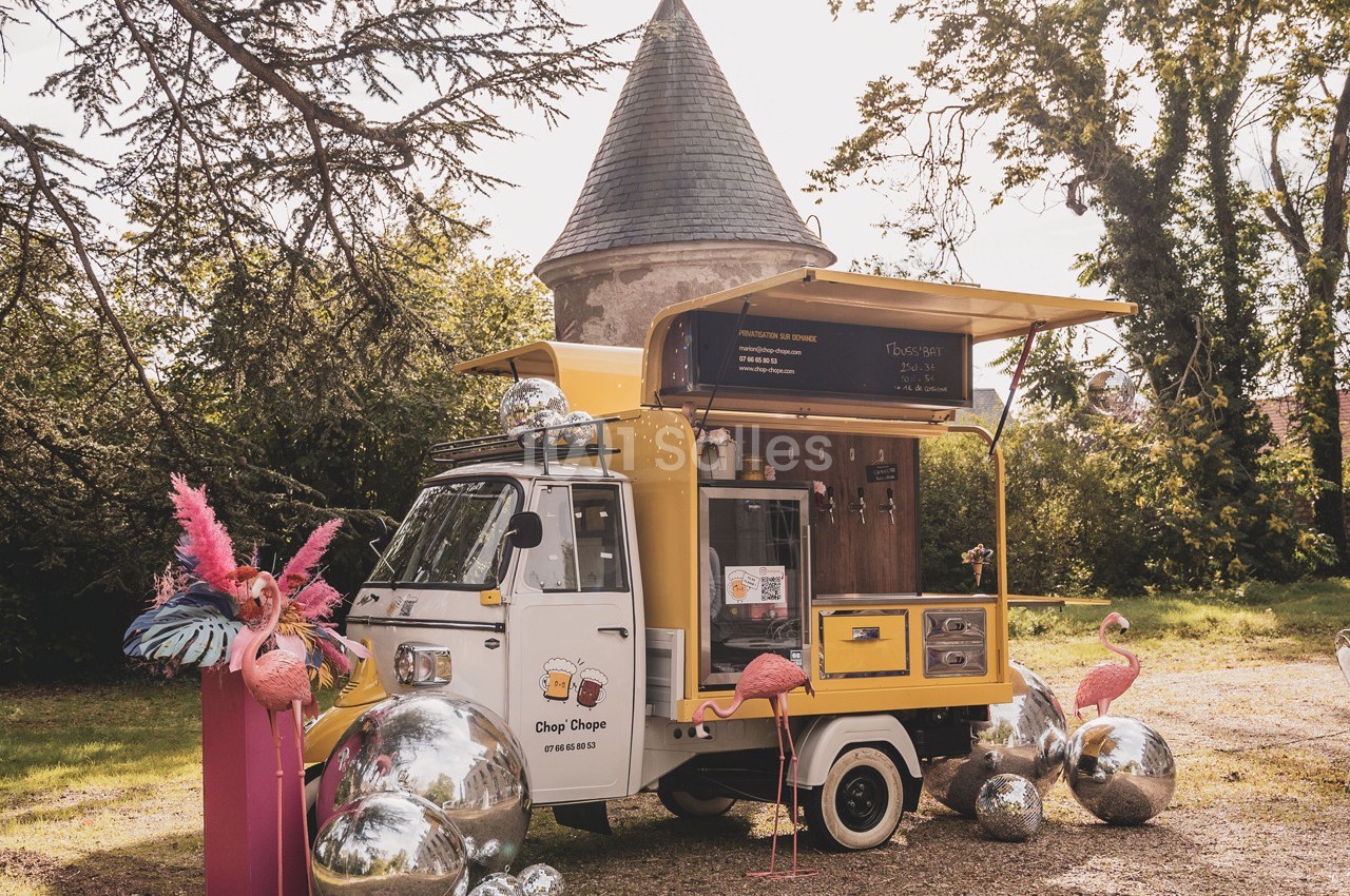 Camion de street food jaune stationné dans un jardin, entouré de flamants roses décoratifs et de sphères argentées.