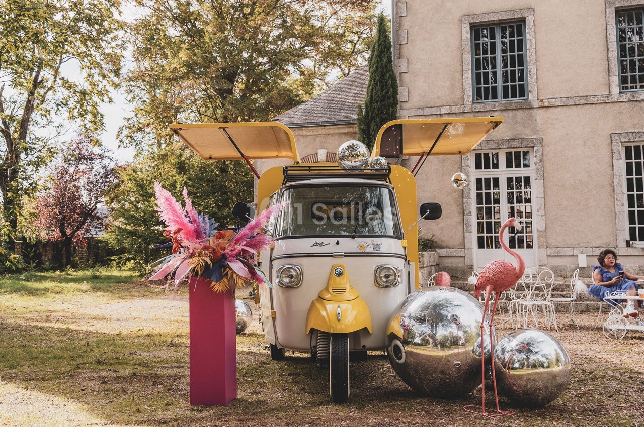 Un triporteur jaune avec ailes ouvertes, entouré de boules miroir, d'un flamant rose et de fleurs colorées, devant un…