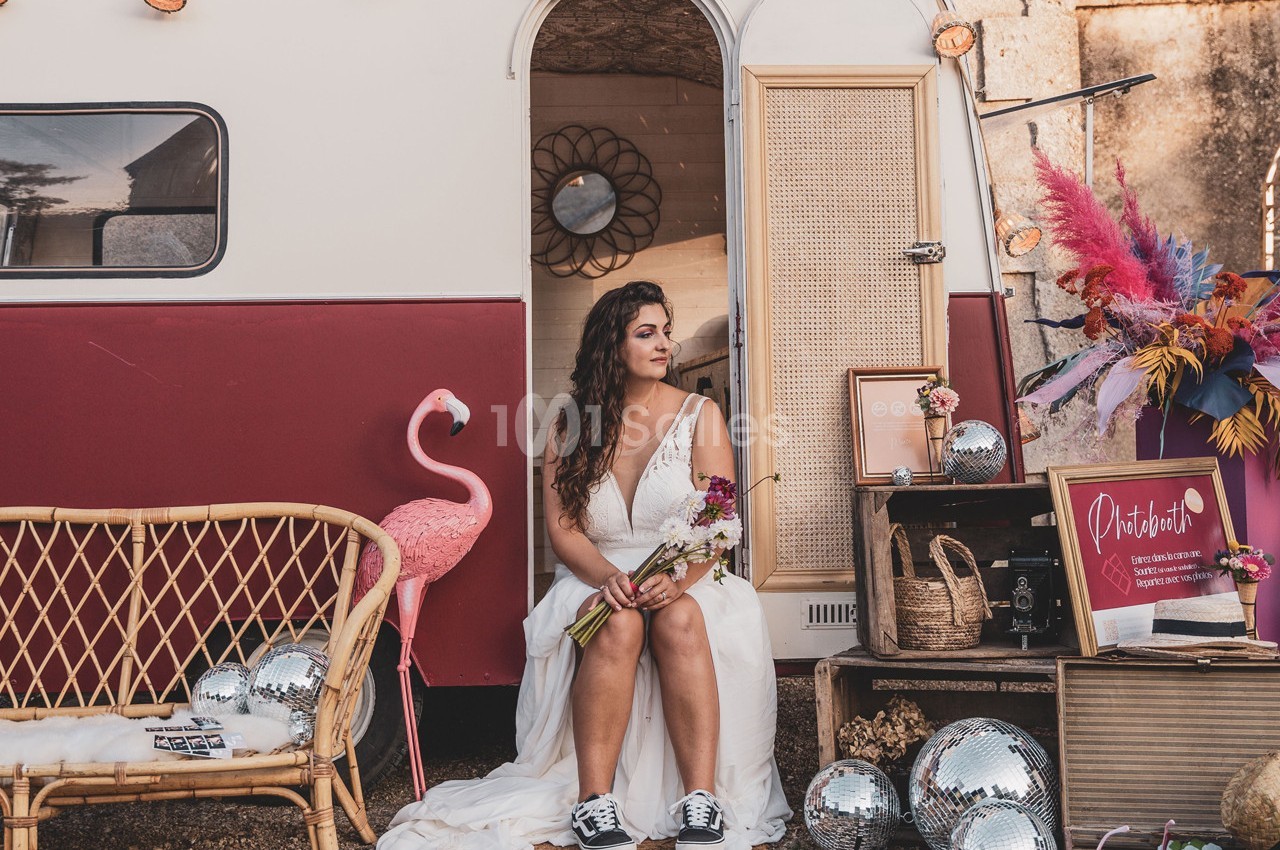 Une femme en robe blanche assise devant une caravane décorée avec des fleurs, des miroirs et des objets rétro.
