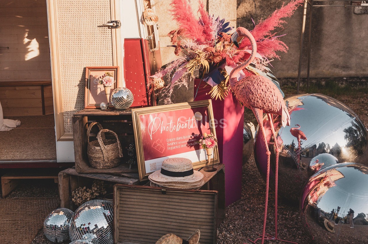 Décor festif avec flamant rose, boules miroir, fleurs colorées et accessoires devant une caravane rétro.