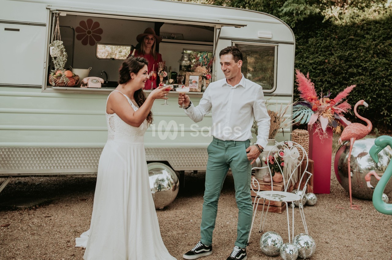 Un couple trinque devant une caravane rétro aménagée, entourée de décorations colorées et festives.