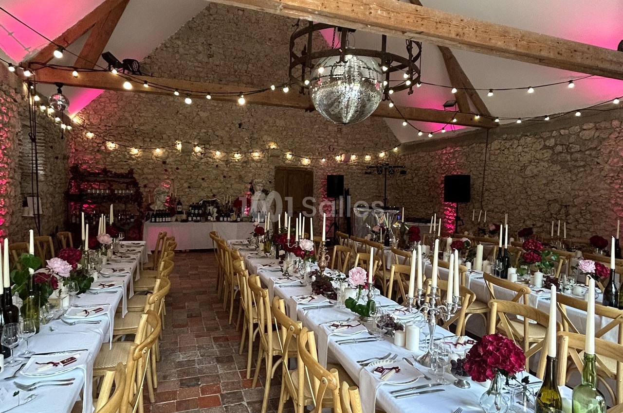 Salle de réception en pierre avec tables décorées, chandeliers, fleurs et guirlandes lumineuses suspendues.