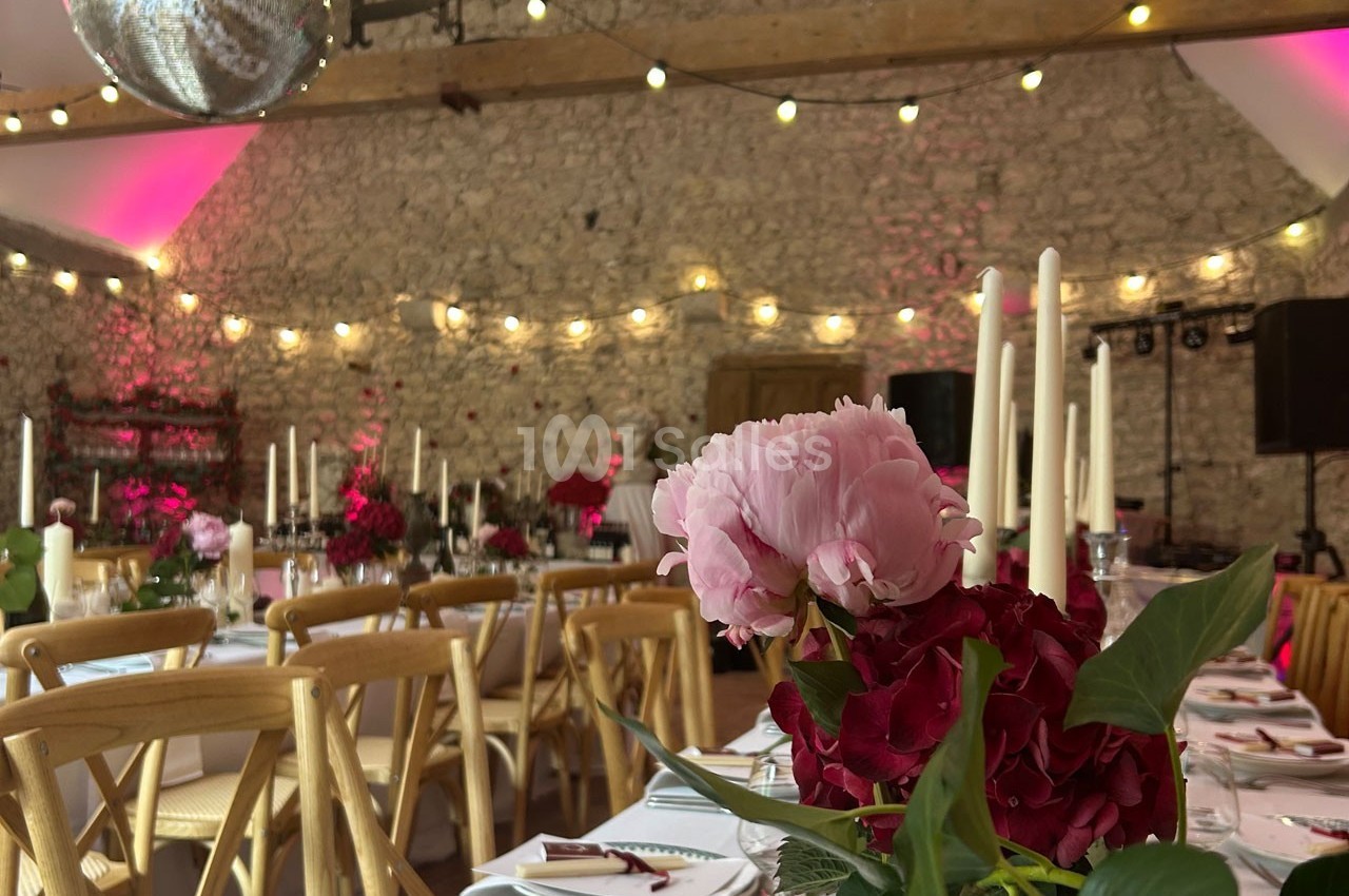 Salle de réception décorée avec des tables dressées, des fleurs roses et rouges, et des guirlandes lumineuses suspendues.