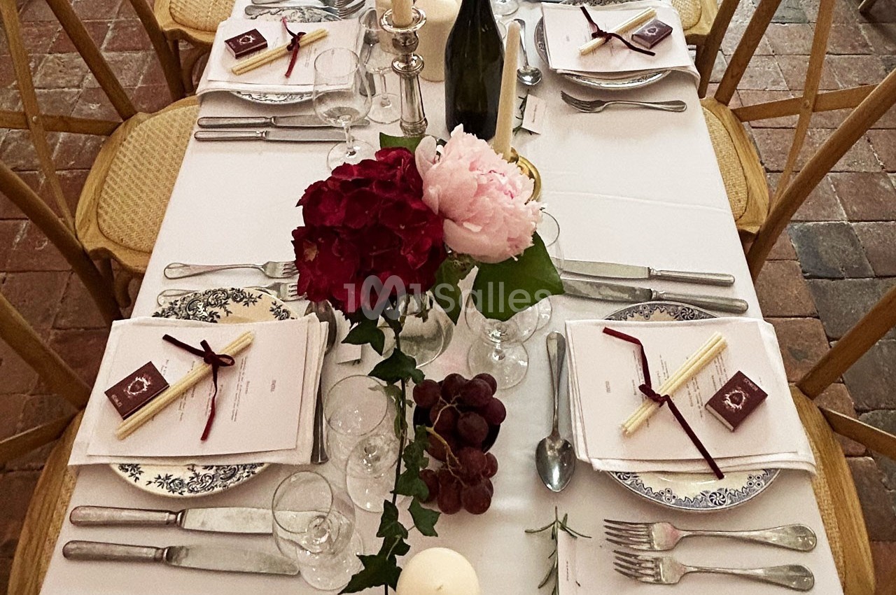 Table dressée avec nappes blanches, vaisselle élégante, fleurs, bougies et grappes de raisin sur un sol en pierre.