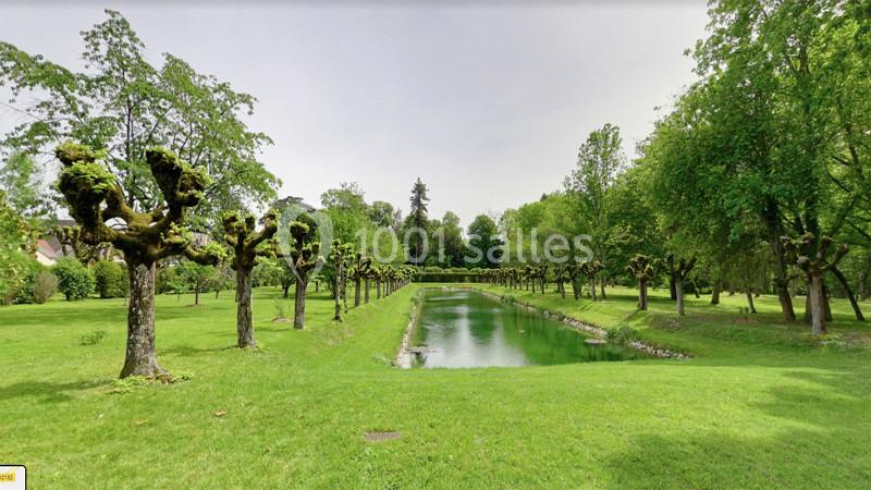 Allée bordée d'arbres taillés, longeant un canal entouré de pelouses et d'arbres dans un parc verdoyant.