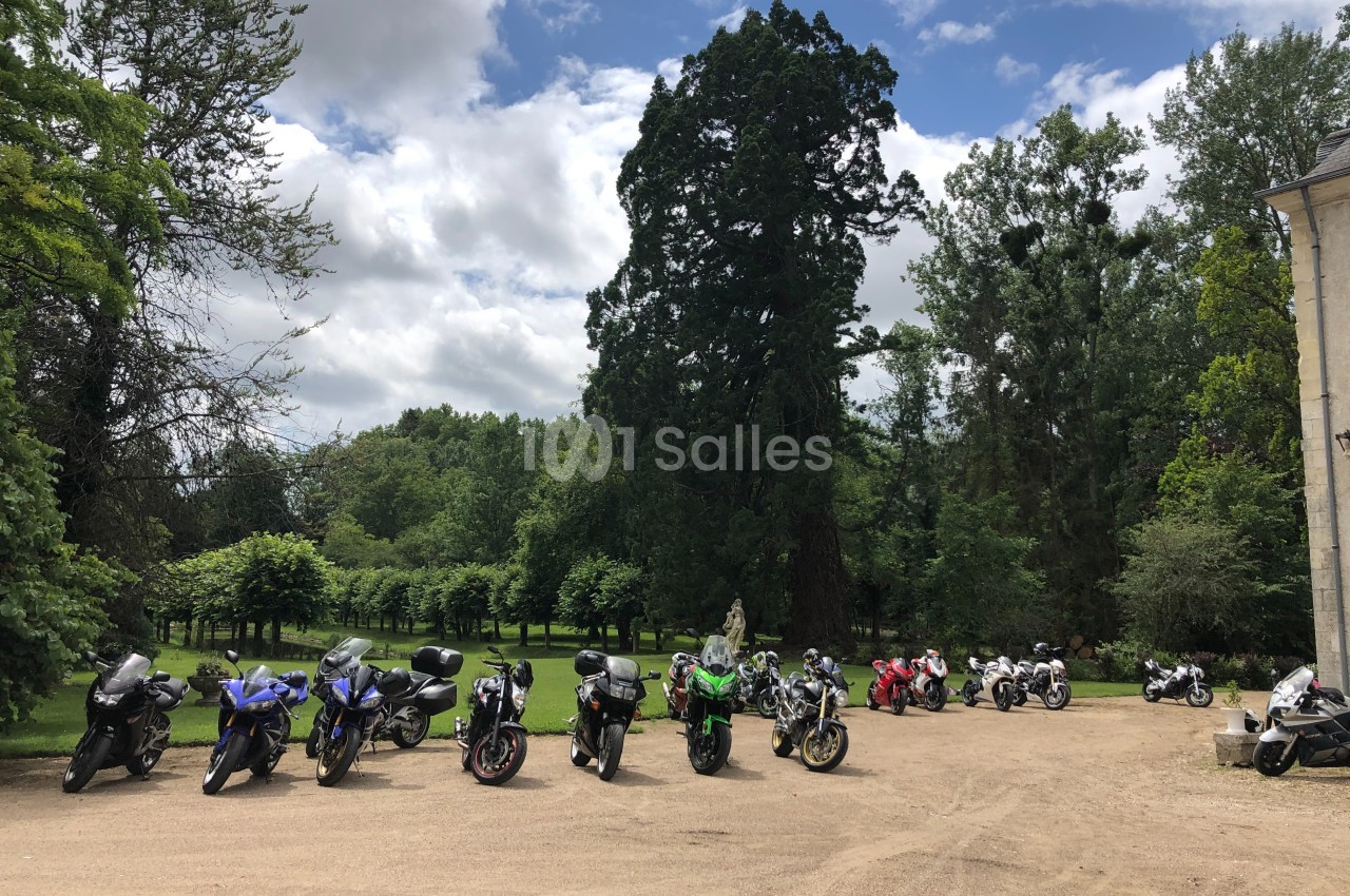 Alignement de motos stationnées sur un chemin en gravier, entouré de verdure et d'arbres sous un ciel partiellement nuageux.