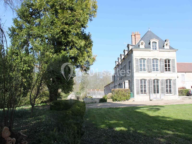 Grande maison en pierre à trois étages avec des volets, entourée d'un jardin arboré sous un ciel dégagé.