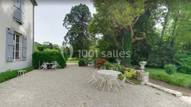 Terrasse en gravier avec tables et chaises en fer forgé, entourée d'arbres et de verdure.