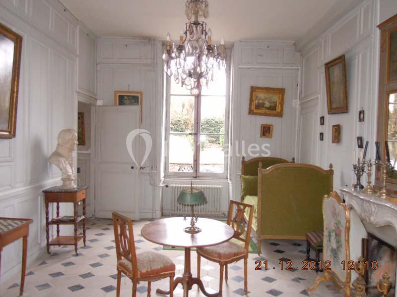 Salle lumineuse avec mobilier ancien, cheminée, lustre, bustes et tableaux, donnant sur une fenêtre avec vue sur jardin.