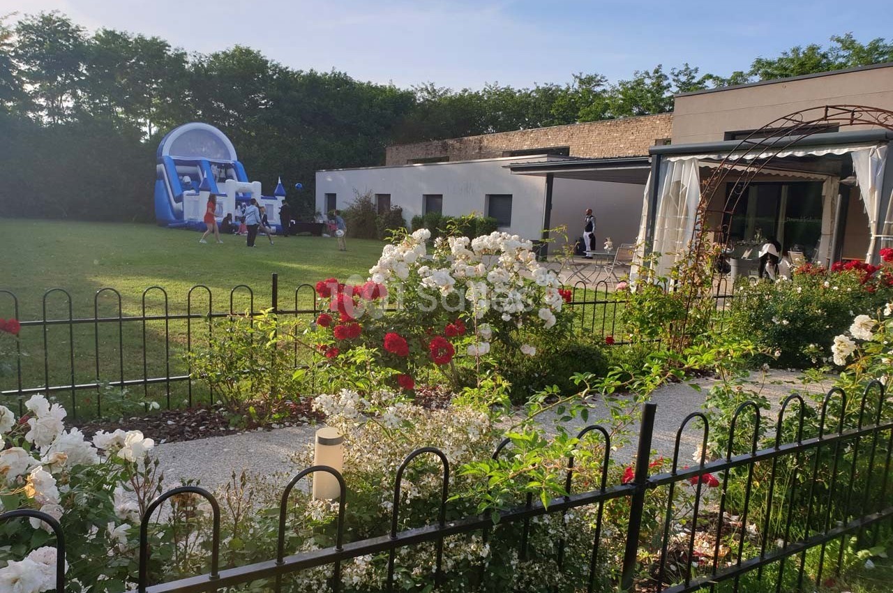 Jardin fleuri avec rosiers, pelouse, structure gonflable en arrière-plan et bâtiment moderne à droite.